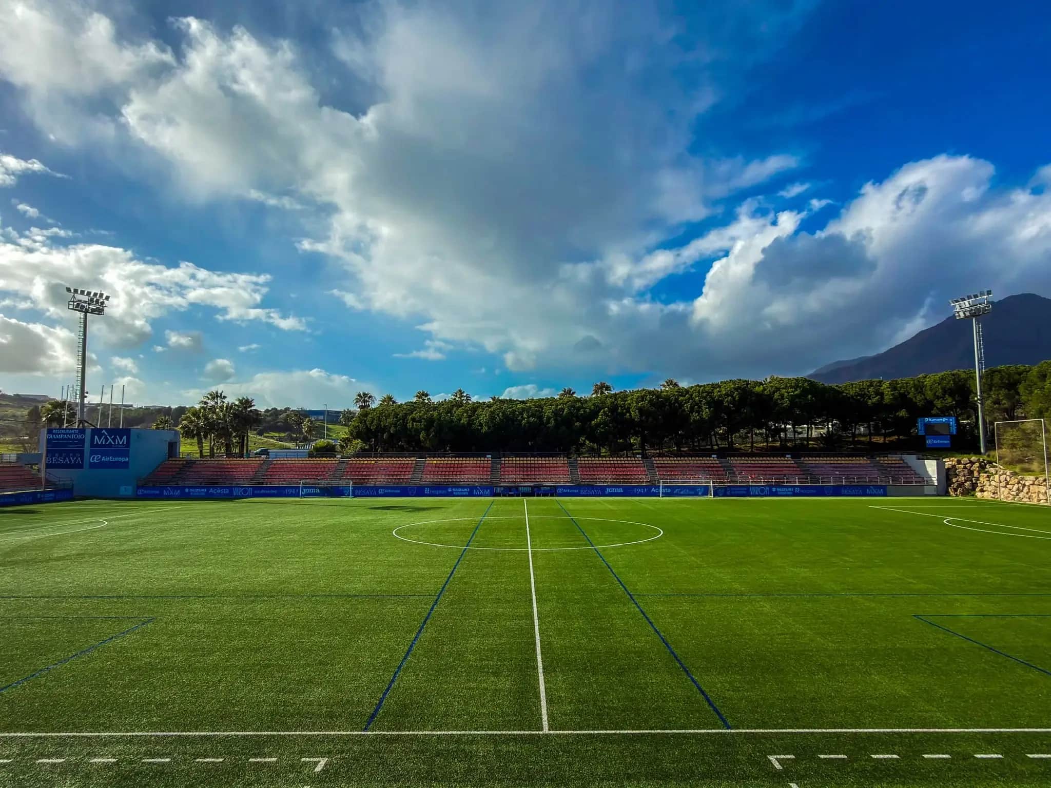 El Estadio Francisco Muñoz Pérez de Estepona, con el césped y la grada vacía bajo un cielo con nubes y la sierra al fondo.